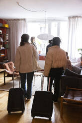 Rear view of female friends standing with wheeled luggage in apartment - MASF41866