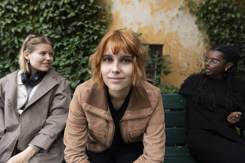 Portrait of blond woman with female friends talking while sitting on bench at park - MASF41878