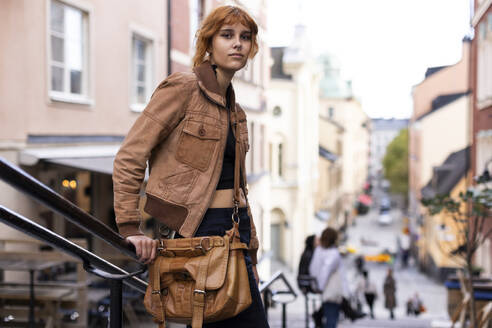 Portrait of young woman with bag and jacket standing near railing in city - MASF41886