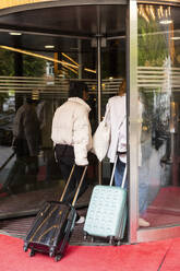 Young female tourists with wheeled luggage entering through revolving door at hotel - MASF41889