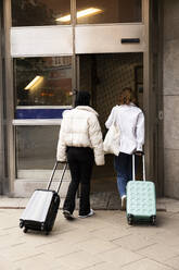 Rear view of young female tourists with wheeled luggage entering in hotel - MASF41890