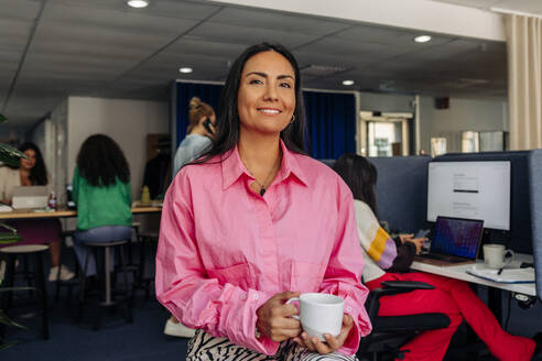 Portrait of smiling female entrepreneur holding coffee cup while sitting at office - MASF41993