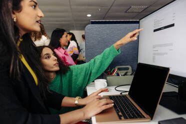 Businesswoman pointing at monitor screen with colleague working on laptop at office - MASF42001