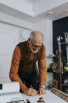 Senior male owner writing on paper at desk while working in antique shop - MASF42017