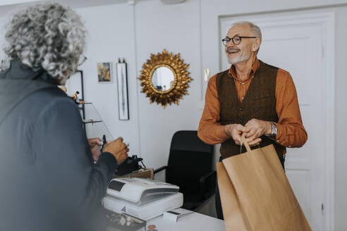 Smiling senior male entrepreneur giving paper bag to female customer while shopping in store - MASF42023