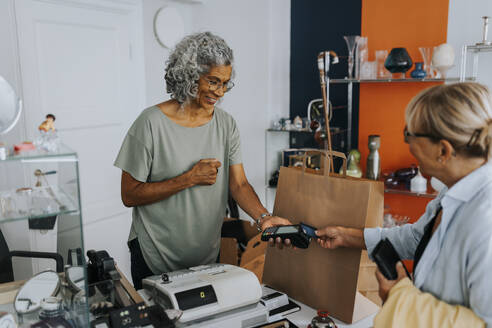 Senior female customer paying through credit card to saleswoman at checkout of antique shop - MASF42033