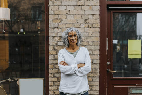 Portrait of smiling senior female entrepreneur with arms crossed standing outside antique shop - MASF42037