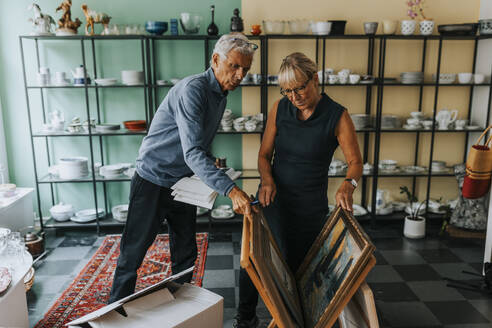 Elderly male and female entrepreneurs examining picture frames in antique shop - MASF42049