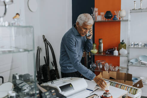 Senior salesman with coffee cup talking on smart phone in antique shop - MASF42058