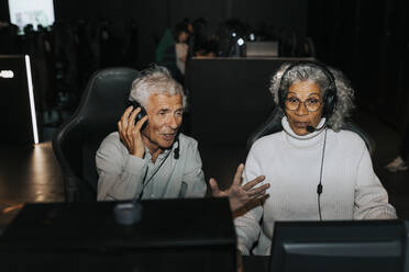 Senior man wearing headset sitting by female friend playing video game on computer at gaming lounge - MASF42078