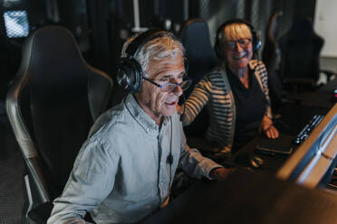 Elderly man with headset playing video game by female friend at gaming center - MASF42114