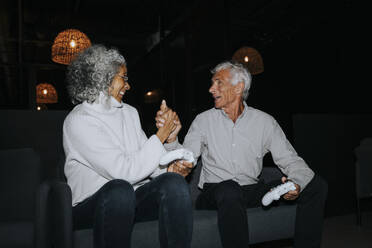 Happy senior couple holding hands while sitting on sofa in gaming center - MASF42120