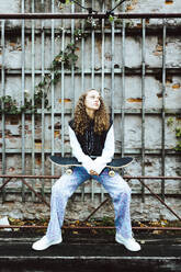 Contemplative girl with skateboard sitting on railing against wall - MASF42127