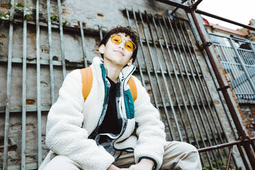 Portrait of boy wearing sunglasses sitting on railing against wall - MASF42133