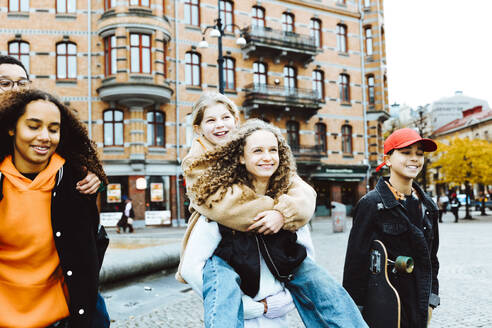 Happy girls giving piggyback ride to male and female friends on footpath in city - MASF42148