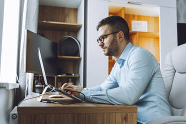 Freelancer wearing eyeglasses and working on laptop at home office - OLRF00069