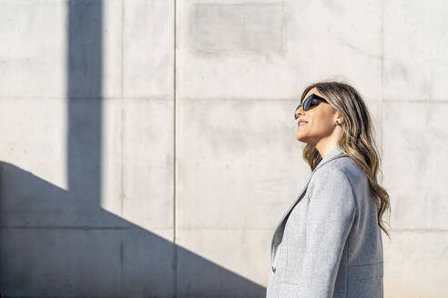 Lächelnde Geschäftsfrau mit Sonnenbrille vor einer Mauer an einem sonnigen Tag - DLTSF03742