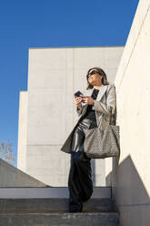 Smiling businesswoman with purse and smart phone leaning on wall - DLTSF03749