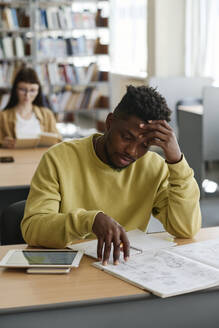 Tired student with head in hand reading book at library - DSHF01516