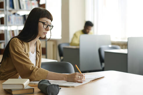 Studentin mit Brille, die in der Bibliothek in ein Buch schreibt - DSHF01519