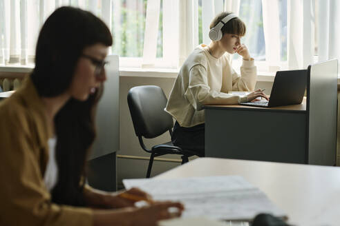 Student wearing wireless headphones and using laptop near window in library - DSHF01520