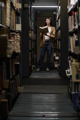 Young student reading book amidst shelves in library - DSHF01529