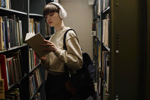 Junge Studentin mit drahtlosen Kopfhörern, die in der Nähe von Regalen ein Buch liest - DSHF01534