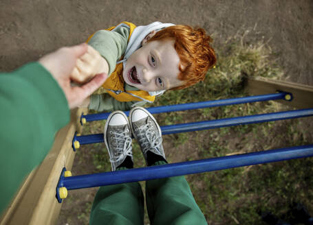 Verspielter Junge hält die Hand seiner Mutter auf einer Leiter auf dem Spielplatz - MBLF00211