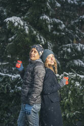 Smiling man and woman standing with cups of punch drink near fir tree - TILF00053