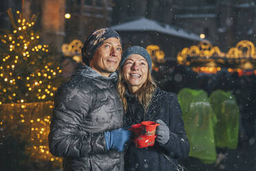 Smiling man and woman holding cups of punch drink at Christmas market - TILF00060