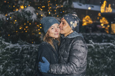 Smiling man kissing woman near illuminated fir tree - TILF00061