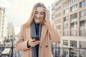 Glückliches blondes Mädchen, das ein Smartphone vor einem Gebäude benutzt - TAMF04049