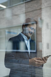 Smiling businessman using mobile phone seen through glass - JOSEF22439