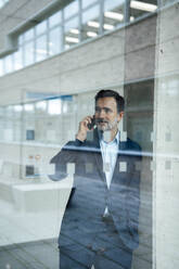 Smiling mature businessman talking on mobile phone seen through glass - JOSEF22443
