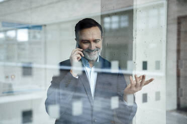 Smiling businessman gesturing and talking on smart phone seen through glass - JOSEF22444