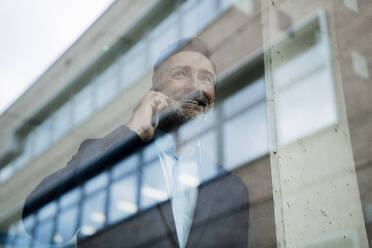 Glücklicher Geschäftsmann, der mit seinem Handy telefoniert und durch ein Fenster schaut - JOSEF22447