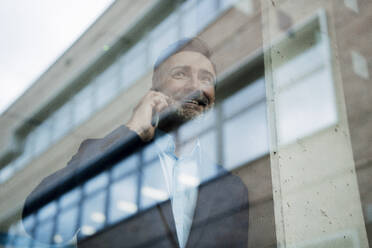 Happy businessman talking on mobile phone and looking through window - JOSEF22447