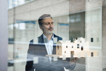 Mature architect holding model house seen through glass - JOSEF22452