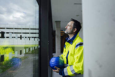 Contemplative engineer standing near window with hardhat in office - JOSEF22459