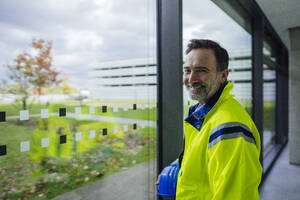 Lächelnder Ingenieur, der einen Schutzhelm trägt und in der Nähe eines Fensters im Büro steht - JOSEF22463