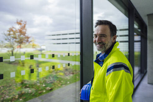 Smiling engineer holding hardhat and standing near window in office - JOSEF22463