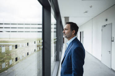 Contemplative businessman looking through glass window in corridor - JOSEF22465