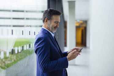 Lächelnder Geschäftsmann, der sein Smartphone in der Nähe eines Glasfensters im Bürokorridor benutzt - JOSEF22468