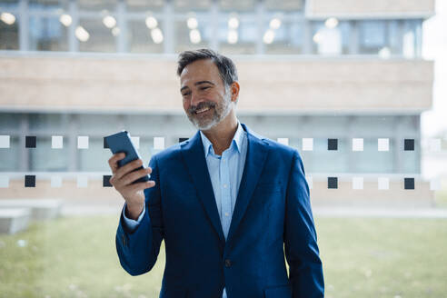 Lächelnder Geschäftsmann, der ein Smartphone vor einer Glasscheibe im Büro benutzt - JOSEF22470