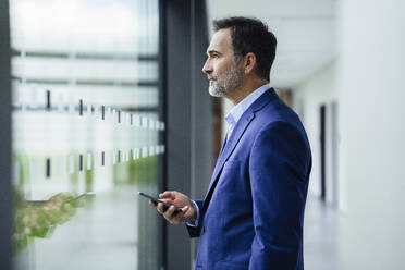 Businessman holding smart phone and looking through window in office corridor - JOSEF22474