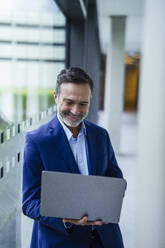 Smiling businessman using laptop near glass window in office - JOSEF22479