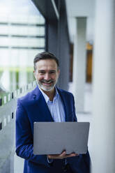 Smiling businessman standing with laptop in office corridor - JOSEF22480