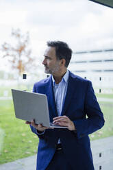 Contemplative businessman holding laptop and looking through glass window in office - JOSEF22481