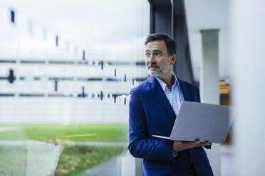 Businessman holding laptop and looking through glass window in office - JOSEF22482