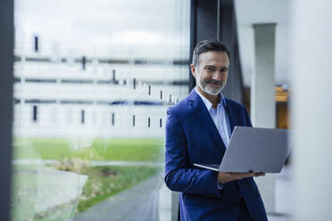 Smiling businessman holding laptop near glass window in office corridor - JOSEF22483